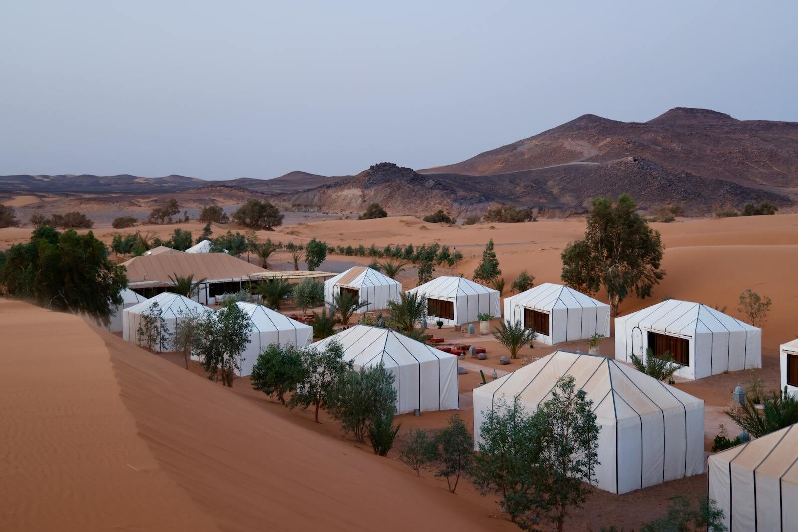 Aerial view of luxury tents in desert landscape with hilly background and twilight ambiance.